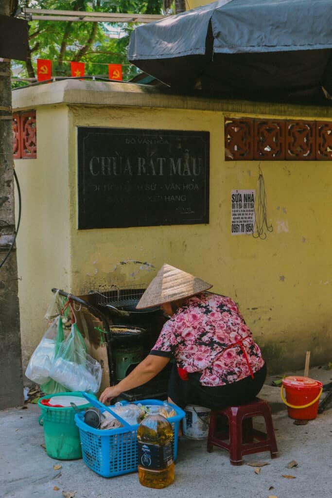 Hanoi street food vendor with conical hat near a temple, a hidden gem in Vietnam's capital.