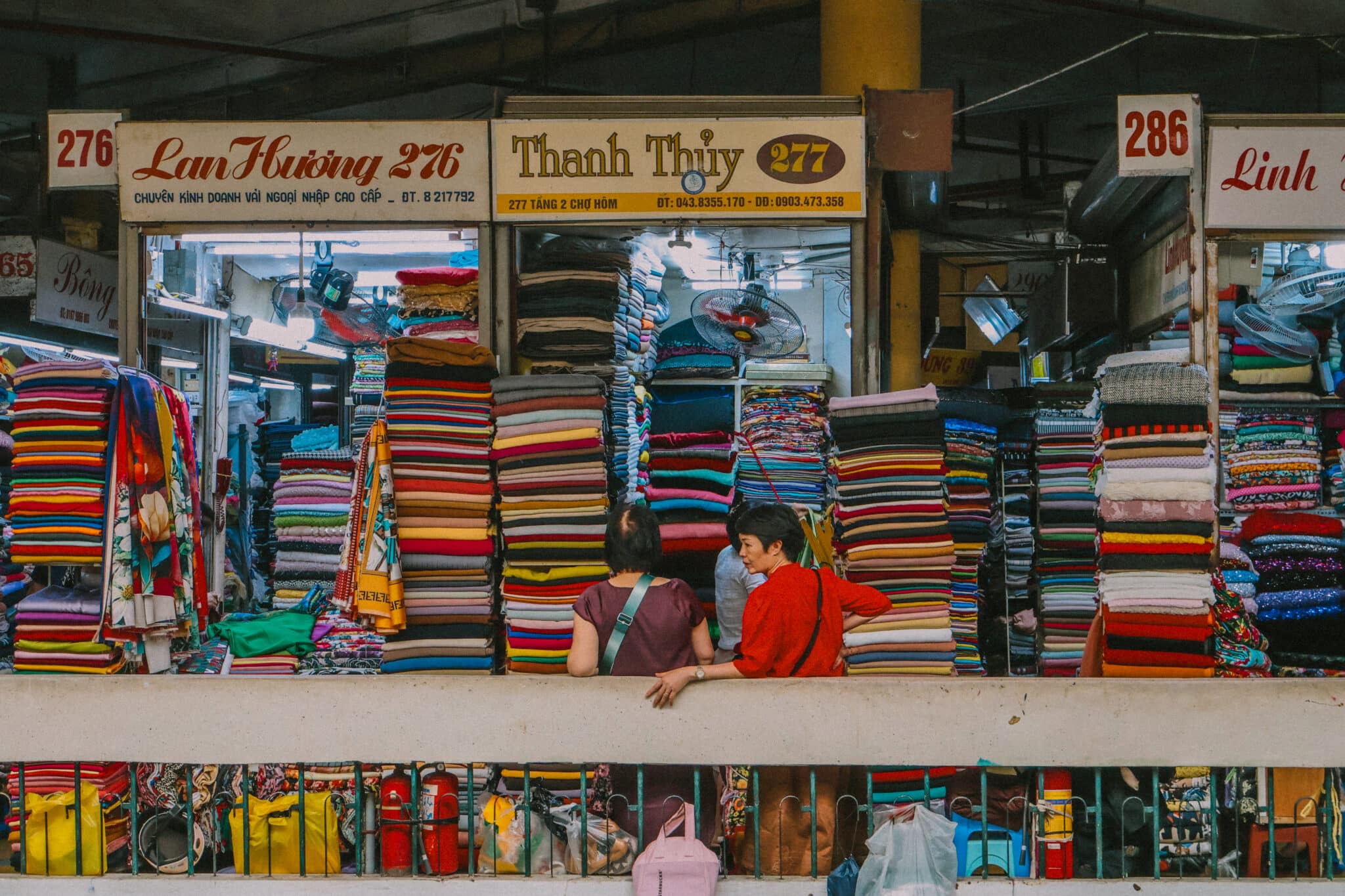 Hanoi Hom market stall with colorful fabric stacks and two women. Things to do in Hanoi: explore local markets.