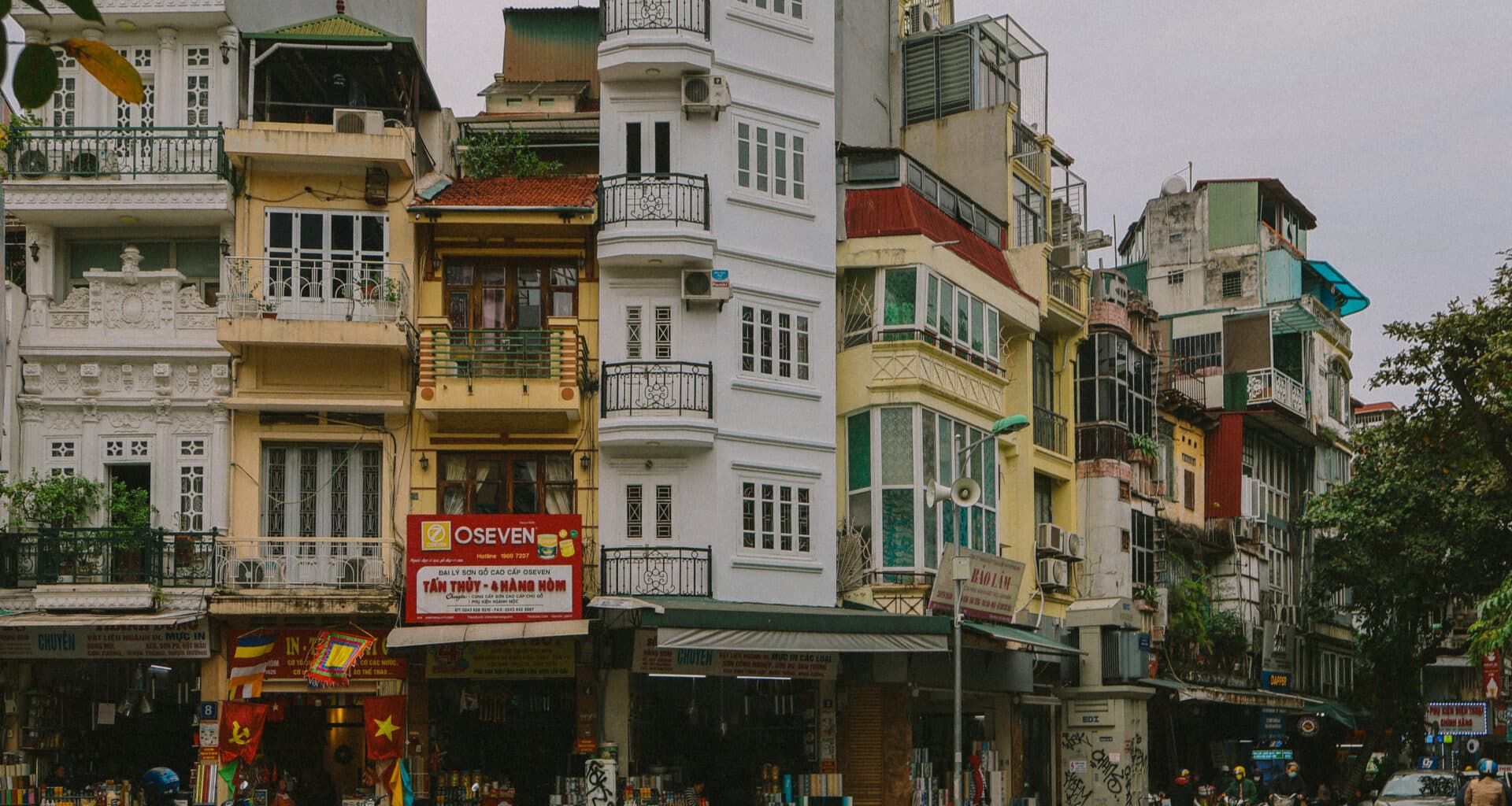 Hanoi street scene with colorful buildings and motorbikes. OSEVEN sign visible.