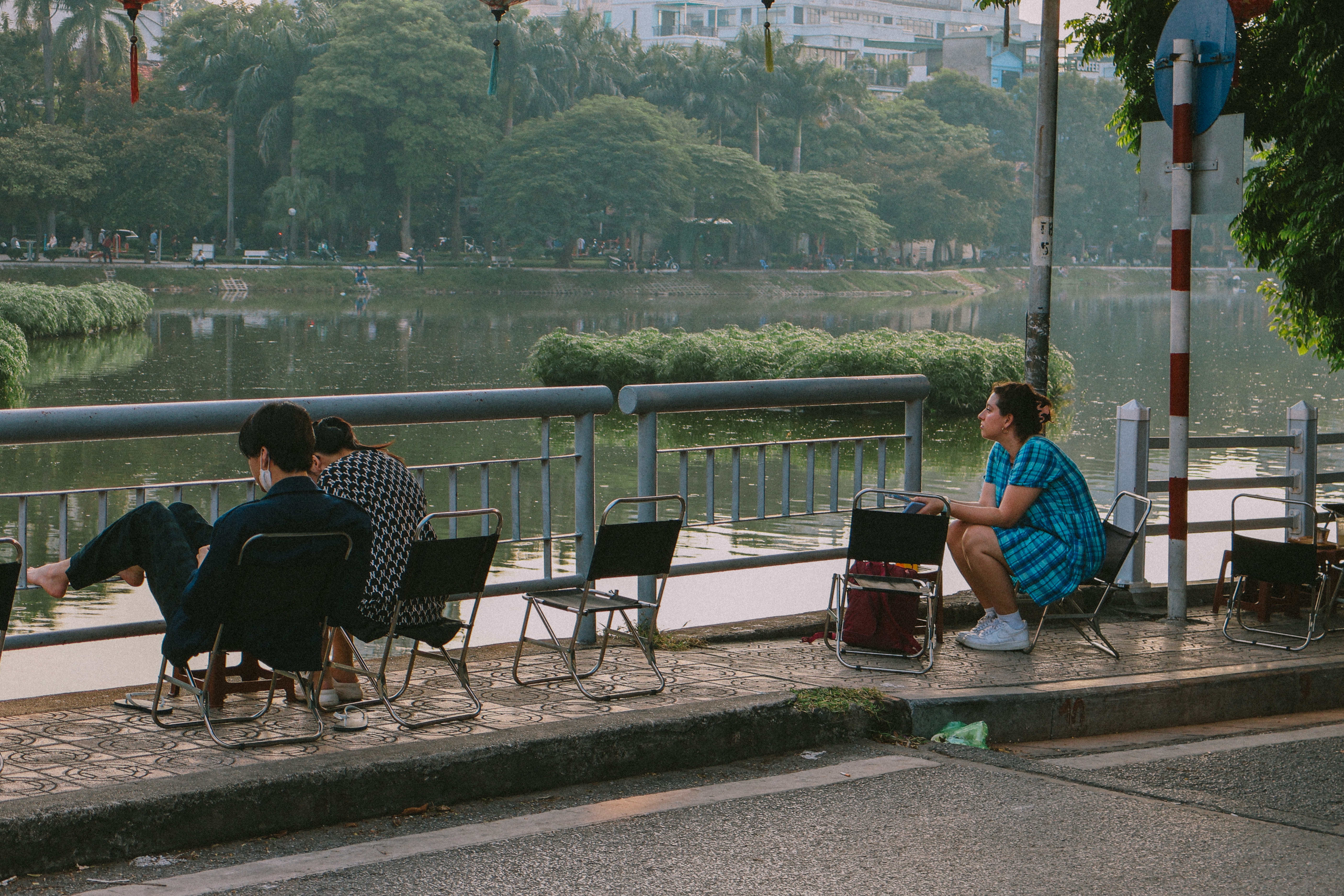People relaxing by a lake in Hanoi, Vietnam, sitting on small chairs. Urban lakeside scene.