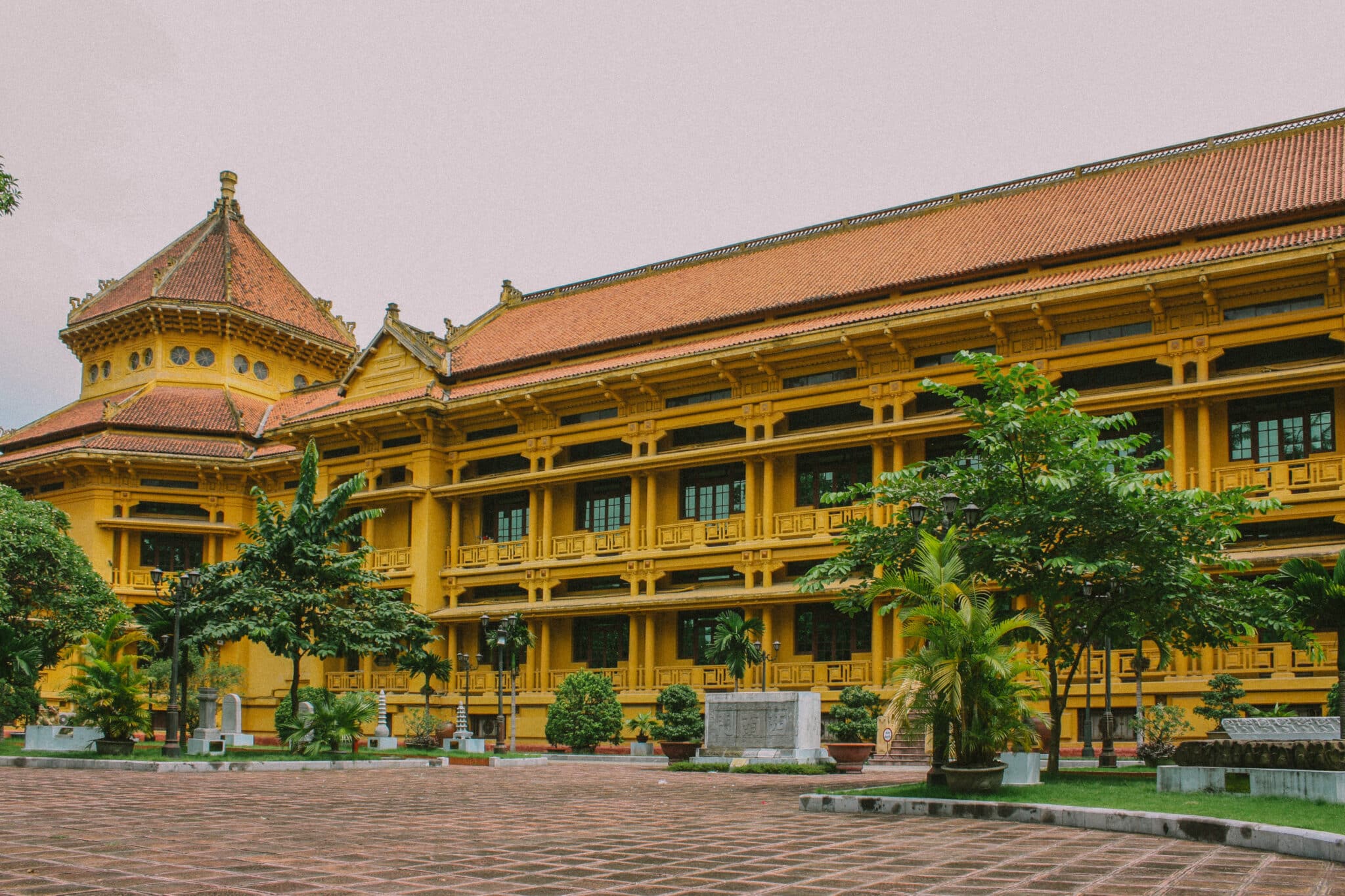 Exterior of a yellow Hanoi museum with a red tile roof, surrounded by trees and a brick courtyard.