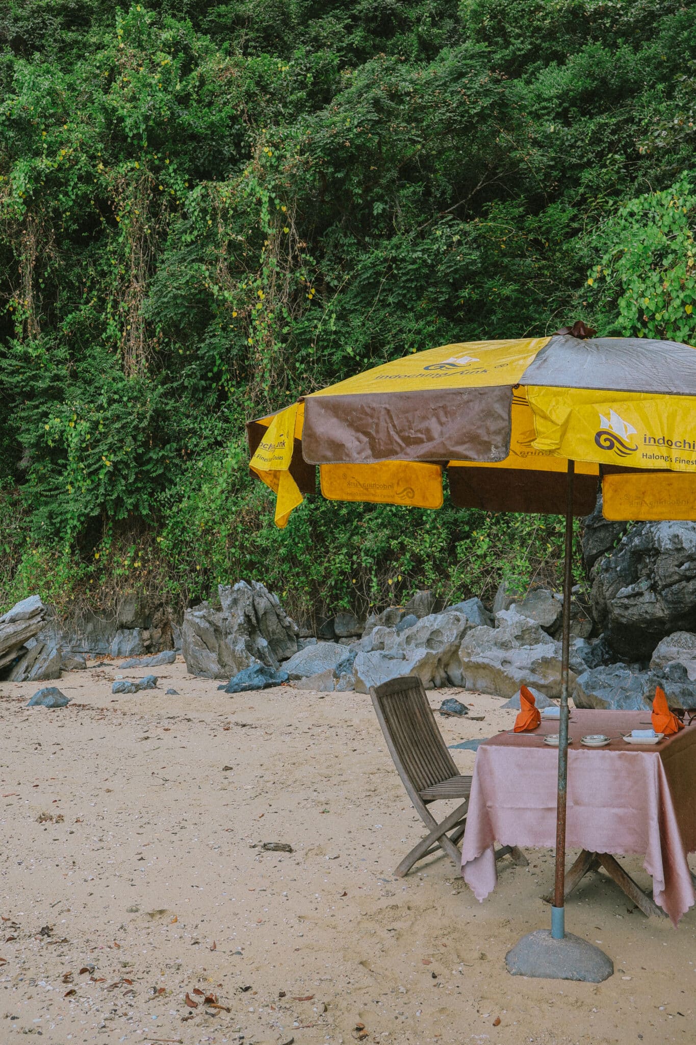 Table set for dining under a yellow umbrella on a Ha Long Bay beach, L'Amour Junk