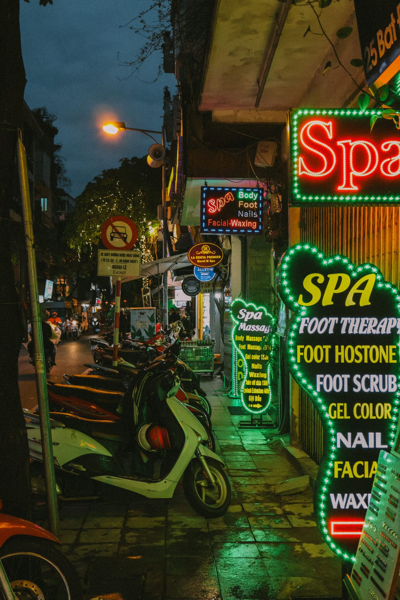 Neon spa signs glow at dusk in Hanoi's Old Quarter, with motorbikes parked nearby.