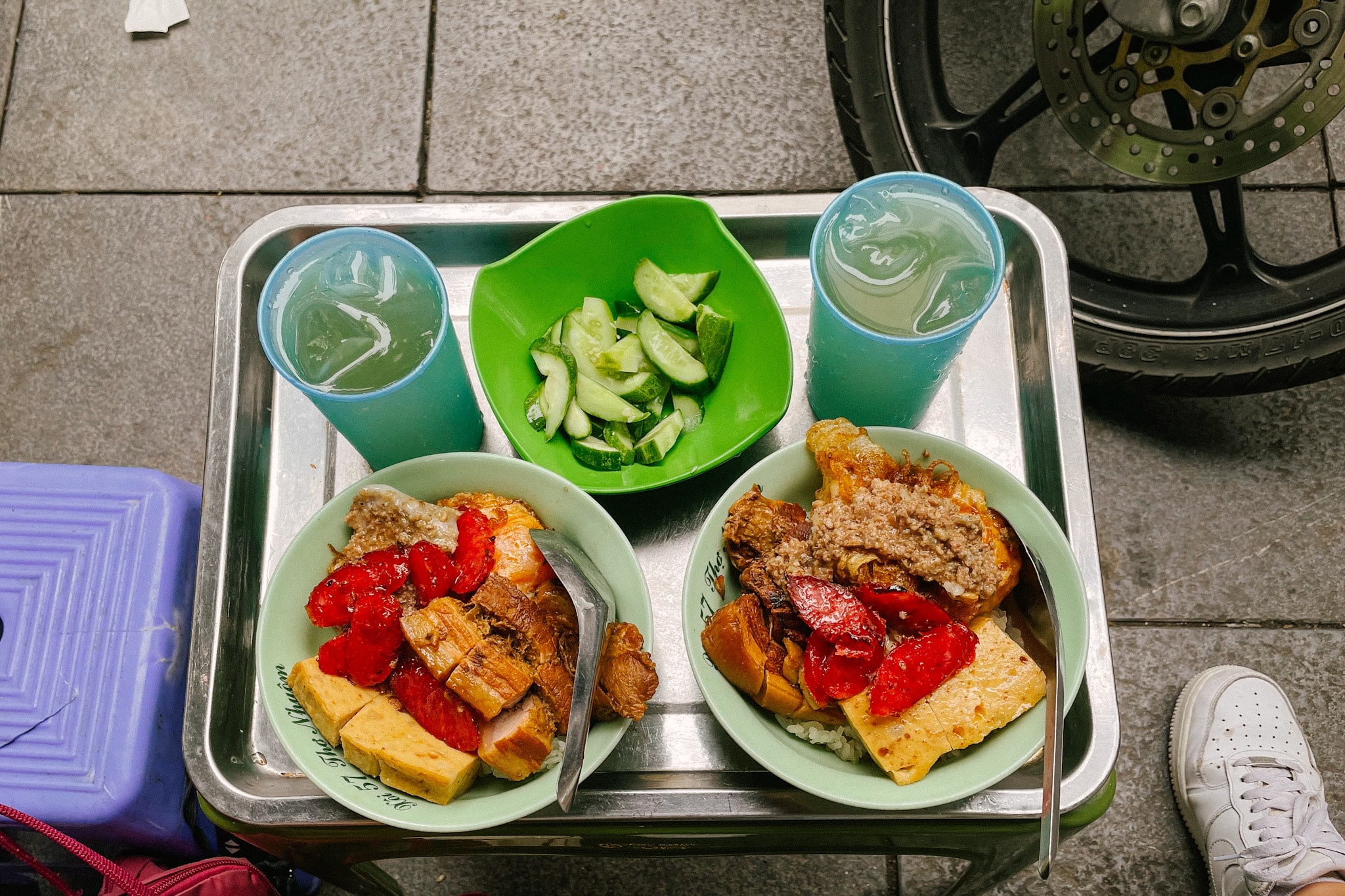 Hanoi street food: two bowls of xoi sticky rice with meat, tofu, and pickled vegetables, and drinks.
