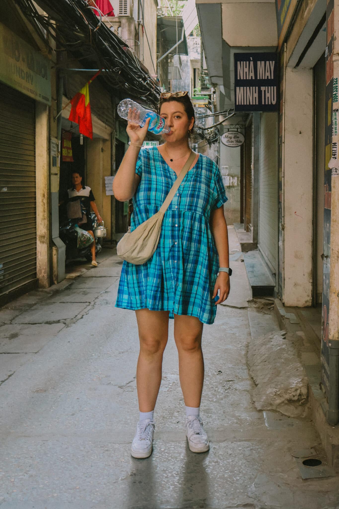 Woman drinking water in a narrow Hanoi street with signs.